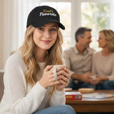Woman wearing a 'Dad's Favorite' cap holding a mug, with a blurred background of two people talking.