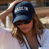 Person wearing a navy blue cap with 'MADE IN Los Angeles' text, sunglasses, and a light pink sweater.