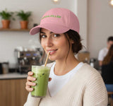 Woman wearing a pink cap with 'Matcha Killer' text, holding a green drink in a kitchen setting.